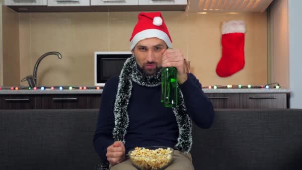 Muž Nový rok Santa Claus klobouk jí popcorn, pít pivo, kouká na televizi, směje se, úsměv — Stock video