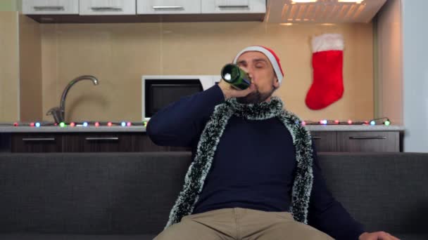 Szalony człowiek w kapeluszu Mikołaja siedzi na kanapie, otwiera butelkę okiem, pije piwo — Wideo stockowe