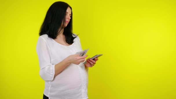 Rica joven embarazada con cabello negro cuenta con dinero en un fondo amarillo — Vídeo de stock