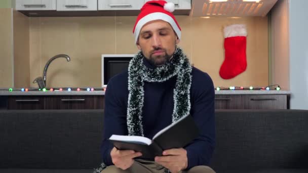 Homem adulto de chapéu vermelho Papai Noel abre caderno, escreve sonhos no próximo ano — Vídeo de Stock