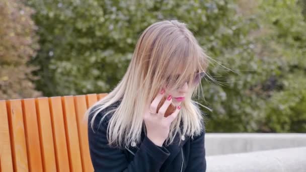 Женщина в солнечных очках разговаривает по мобильному телефону, сидит на скамейке в открытом парке — стоковое видео