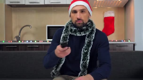 Man Nieuwjaar Kerstman hoed gaat op de bank zitten, neemt afstandsbediening, zet TV aan — Stockvideo