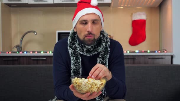 Mężczyzna w Nowym Roku Santa Claus kapelusz je popcorn, ogląda telewizję na tle kuchni — Wideo stockowe