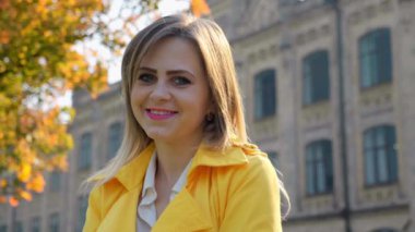 Beyaz bluzlu, sarı pelerinli kadın gülümseyip dışarıdaki kameraya bakıyor.