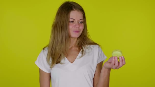 Frau im weißen T-Shirt hält eine gelbe Makrone in der Hand, lächelt und zeigt wie — Stockvideo