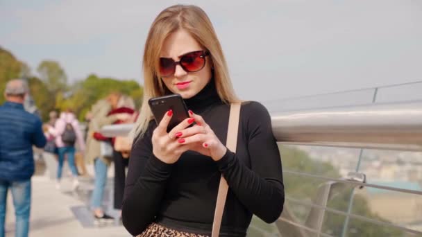 Mujer en gafas de sol y cuello alto negro utiliza teléfono inteligente al aire libre — Vídeos de Stock