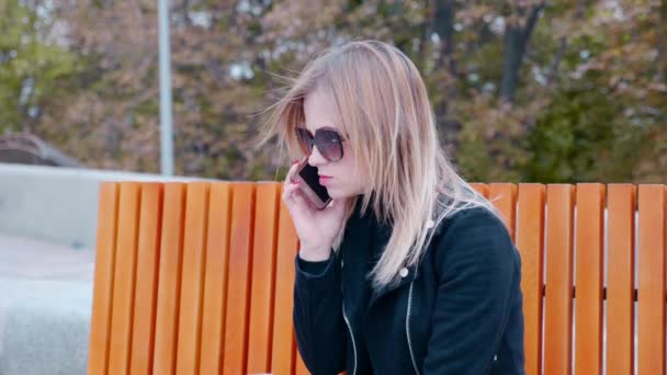 Mujer con gafas de sol y chaqueta negra sostiene teléfono inteligente cerca de la oreja al aire libre — Vídeos de Stock