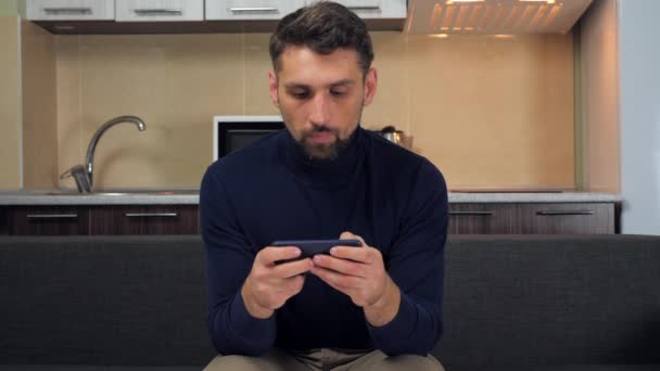 Przystojny mężczyzna gracz siedzi na kanapie w domu grając w gry wideo na smartfonie — Wideo stockowe