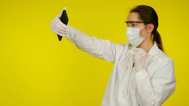 Koruyucu tıbbi maskeli, gözlüklü, eldivenli doktor akıllı telefondan selfie çekiyor.