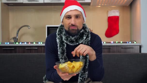 Glad man i röd jultomte hatt äter potatischips, tittar på TV, njuter, ler — Stockvideo