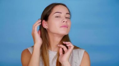 Gri tişörtlü kadın kameraya poz veriyor, saçlarını mavi arka planda düzleştiriyor.