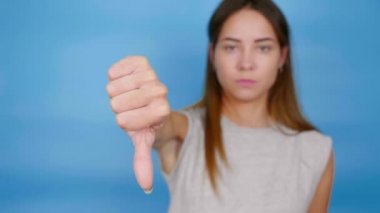 Gri tişörtlü ciddi bir kadın mavi arka planda sevilmeyen baş parmağını aşağı gösteriyor.