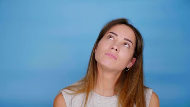 Beautiful woman in gray t-shirt looks up and thinks on blue background — Stock Video