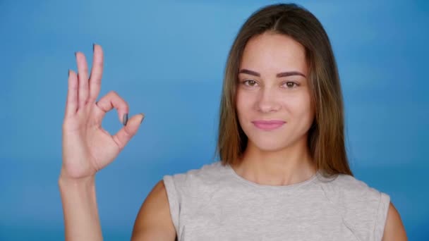 Mooie vrouw in grijs t-shirt glimlacht, heft de hand, toont ok op blauwe achtergrond — Stockvideo