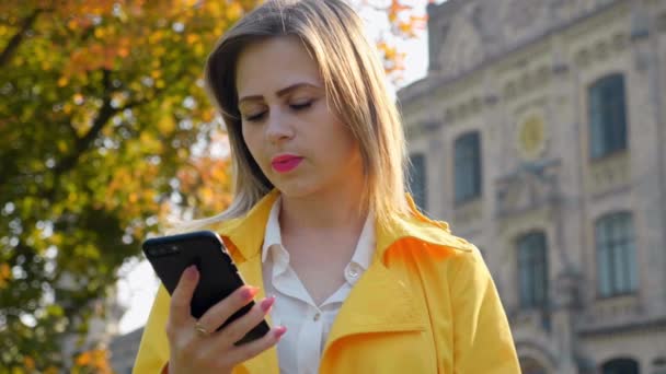 Femme en chemisier blanc et manteau jaune utilise smartphone à l'extérieur. Mouvement lent — Video