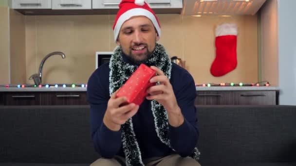 Человек в шляпе Санта-Клауса, ловит красную коробку с подарком в руках, исследует, улыбается — стоковое видео