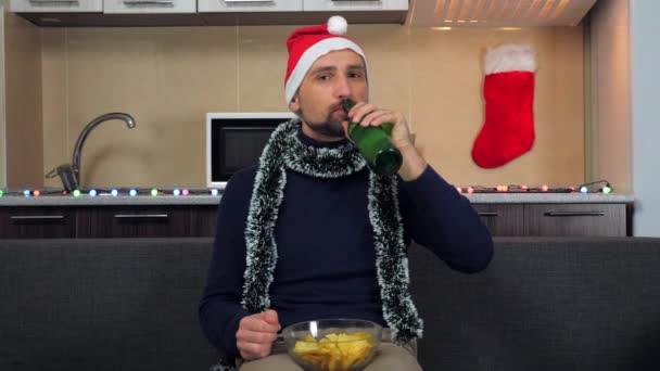 Homem positivo Papai Noel chapéu come batata frita, bebe cerveja, assiste TV, sorri — Vídeo de Stock