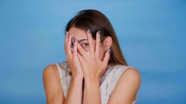 Gri tişörtlü korkak kadın korkuyor, mavi arka planda avuçlarıyla gözlerini kapatıyor.
