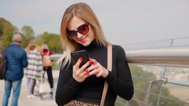 Mujer positiva en gafas de sol, sonrisas de cuello alto negro, utiliza teléfono inteligente al aire libre — Vídeos de Stock