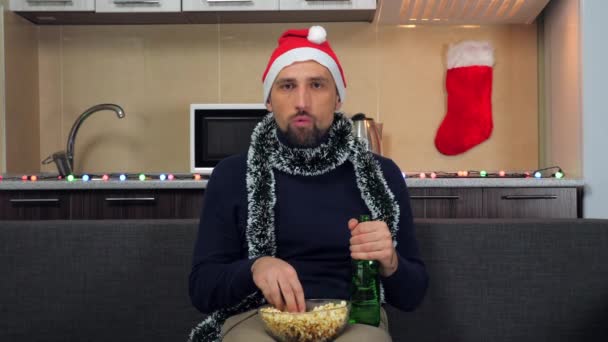 Skoncentrowany człowiek Nowy Rok Kapelusz Świętego Mikołaja je popcorn, pije piwo, ogląda telewizję — Wideo stockowe