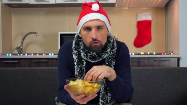 Concentrated man in red Santa Claus hat eats potato chips, watches TV, worries — Stock Video