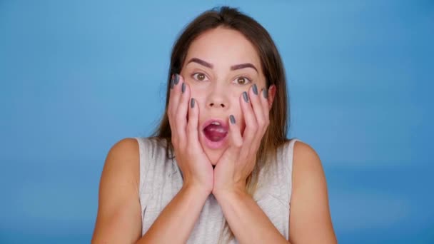 Шокована жінка відкриває рот і кладе долоні на щоки на синьому фоні — стокове відео