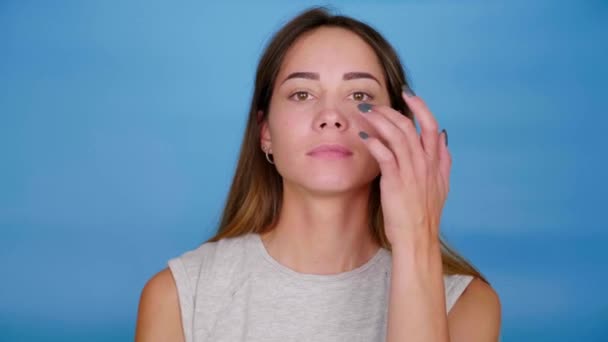Mulher bonita em cinza T-shirt arranhões perto do olho no fundo azul — Vídeo de Stock