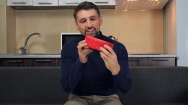 Mavi kazaklı mutlu adam kırmızı kutuyu inceliyor, sarılıyor, hediyesini veriyor, gülümsüyor.