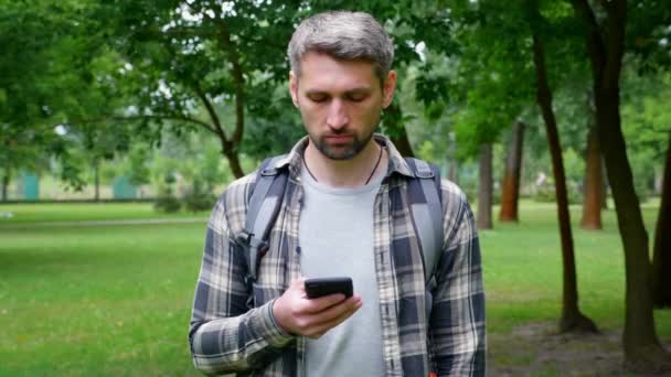 Gris de pelo serio hombre turista stands en parque utiliza teléfono inteligente toque pantalla táctil — Vídeos de Stock