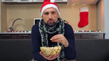 Yoğunlaşmış adam yeni yıl kırmızı Noel Baba şapkası yiyor, patlamış mısır yiyor, kızgın televizyon seyrediyor.