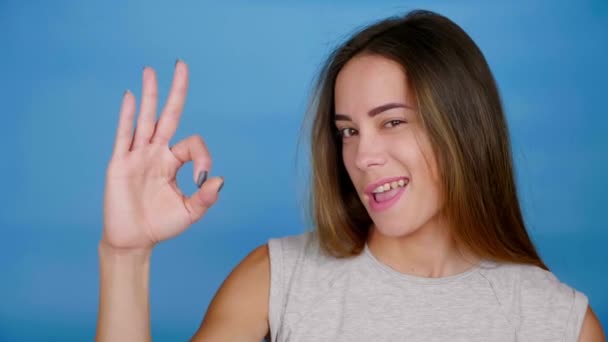 Vacker kvinna höjer handen, visar ok, blinkar ett öga, ler på blå bakgrund — Stockvideo