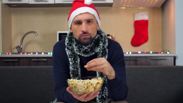 Сконцентрированный человек Новый год красный Санта-Клаус шляпа ест попкорн, сердитый смотреть телевизор — стоковое видео