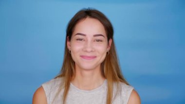 Gri tişörtlü neşeli kadın kameraya bakıyor, poz veriyor, mavi arka planda gülümsüyor.