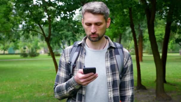 Hombre serio turista de pelo gris se para en el parque utiliza la pantalla táctil del grifo del teléfono inteligente — Vídeos de Stock