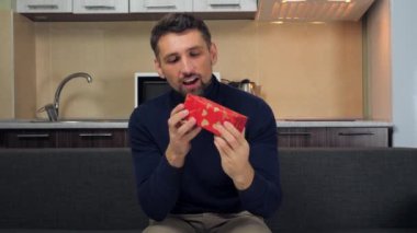 Mavi kazaklı mutlu adam kırmızı kutuyu inceliyor, sarılıyor, hediyesini veriyor, gülümsüyor.
