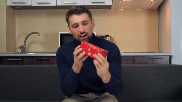 Hombre guapo feliz en jersey azul examina caja roja, abrazos, regocija regalo, sonrisas — Vídeos de Stock