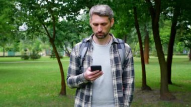 Gri saçlı ciddi turist parkta duruyor. Akıllı telefon dokunuşu ekranı kullanıyor.