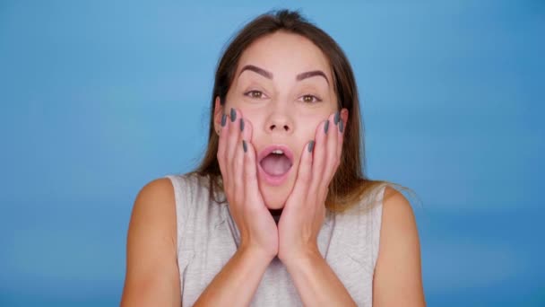 Шокована жінка відкриває рот і кладе долоні на щоки на синьому фоні — стокове відео