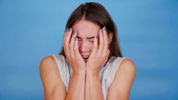 Przestraszona kobieta w szarej koszulce boi się, zamyka oczy dłońmi na niebieskim tle — Wideo stockowe