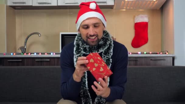 Позитивный человек в новогодней шляпе Санта Клауса, рассматривает коробку с подарком, обнимает, улыбается — стоковое видео