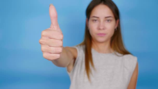Ładna kobieta w szarym t-shircie pokazuje kciuk do góry, jak na niebieskim tle — Wideo stockowe