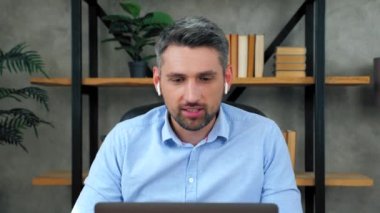Kablosuz kulaklıklı gülümseyen iş adamı çalışanlara video konferansı düzenliyor, internet ofisinde çalışıyor, dizüstü bilgisayar kullanıyor. Erkek öğrenci web kamerasında öğretmenle konuşuyor, uzaktan eğitim.
