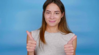 Gri tişörtlü güzel, pozitif kadın parmaklarını yukarı kaldırıyor ve mavi arka planda fotokopi çekerek gülümsüyor. 4K yavaş çekim görüntüleri