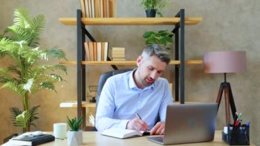 Ev ofisinin çalışmasında gülümseyen adam öğretmenle uzaktan video konferansı, uzaktan eğitim. Kablosuz kulaklıklı sakallı adam dizüstü bilgisayara bakar ve bilgi defteri yazar.