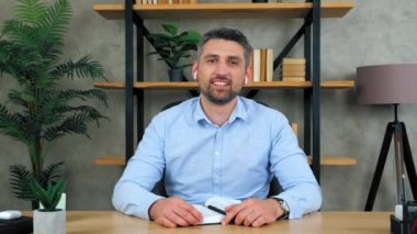 Ofisteki kablosuz kulaklıklarla gülümseyen iş adamı kamerayı selamlıyor kafa sallıyor sohbet konferansı ve web kamerası konseptiyle şirkette potansiyel bir iş görüşmesini yönetiyor.