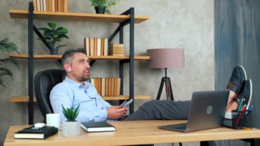 Ofisteki başarılı, güler yüzlü iş adamı kablosuz kulaklık takıyor. Sandalyeye oturuyor, bacakları masada oturuyor. Paranın nereye harcayacağını seçtiğini düşünüyor. 4K yavaş çekim görüntüleri