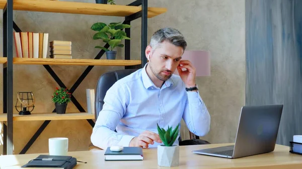 Бизнесмен в домашнем офисе надевает беспроводные наушники с помощью ноутбука — стоковое фото