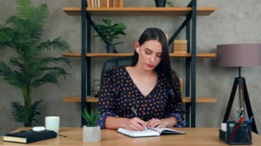 Kendine güvenen güzel esmer iş kadını yönetici CEO 'su ofisteki koltukta oturuyor kablosuz kulaklık takıyor müzik bilgi notaları yazıyor. İngilizce ödevi yapan kadın