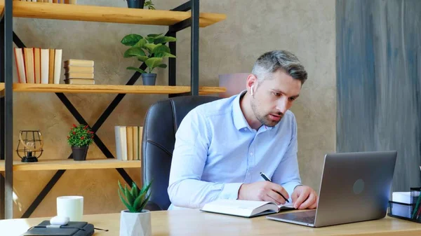 Человек в домашнем офисе в беспроводных наушниках пишет ноутбук выглядит ноутбук компьютер — стоковое фото
