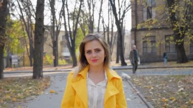 Sarı pelerinli beyaz bluzlu mutlu kadın parkta kameraya bakarak gülümsüyor. 4K yavaş çekim görüntüleri